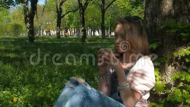 公园里的女孩喝咖啡。 一个十几岁的女孩穿着牛仔裤西装坐在树下。 那个悲伤的女孩喝咖啡。